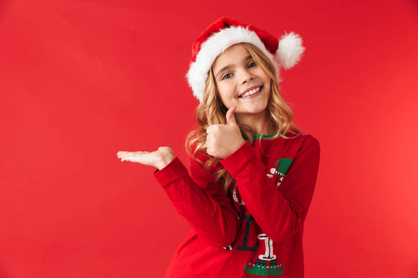 Cheerful little girl wearing Christmas costume standing isolated over red background, presenting copy space - Fotó, kép