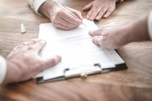 man signing contract at wooden desk with other person pointing at document - Фото, зображення