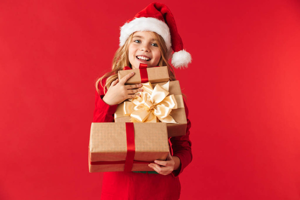 Cheerful little girl wearing Christmas costume standing isolated over red background, holding gift boxes - Fotó, kép