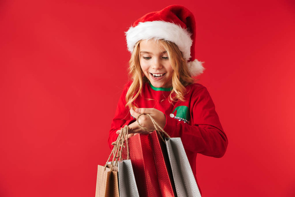 Cheerful little girl wearing Christmas hat standing isolated over red background, carrying shopping bags - Foto, Imagem