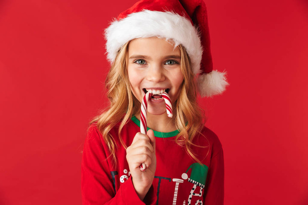 Весела маленька дівчинка в різдвяному костюмі стоїть ізольовано на червоному тлі, їсть різдвяні цукерки з конусами
 - Фото, зображення