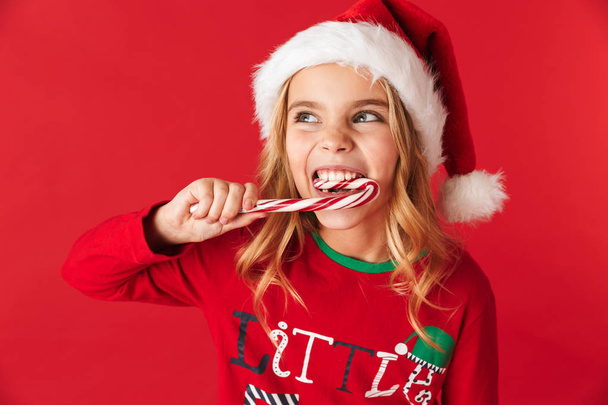 Cheerful little girl wearing Christmas costume standing isolated over red background, eating Christmas candy cone candy - Фото, зображення