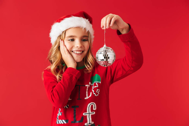 Cheerful little girl wearing Christmas costume standing isolated over red background, holding Christmas tree toy - Photo, image