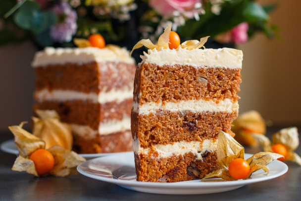 Duas fatias de bolo de cenoura caseiro com nozes, peras e creme de queijo e physalis como decoração nos pratos com uma cesta de flores frescas no fundo - Foto, Imagem