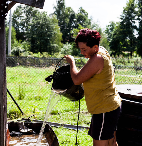 W średnim wieku kobieta napełniania wanny metalowe z naturalnej wody ze studni. Pracowity ogrodnik zbieranie wody przygotowanie do pora sucha. Codziennie chore gospodarstwa domowego. Scena obszar wiejski. - Zdjęcie, obraz