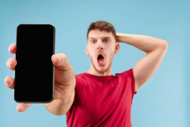 Young handsome man showing smartphone screen over blue background with a surprise face. Human emotions, facial expression concept. Trendy colors - Valokuva, kuva