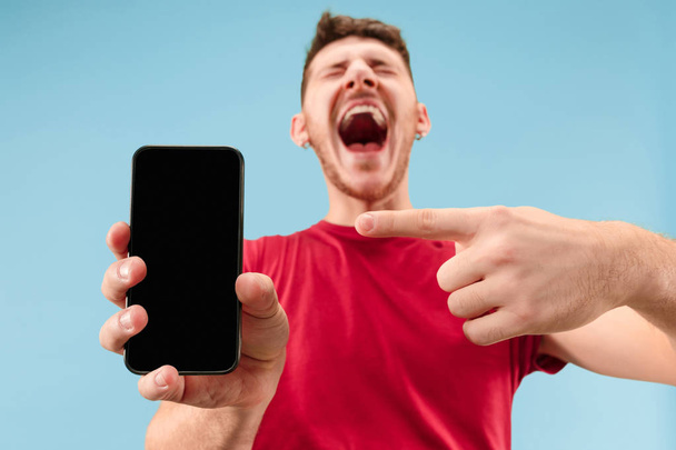 Young handsome man showing smartphone screen isolated on blue background in shock with a surprise face - Foto, Bild