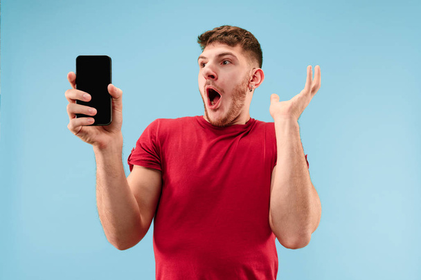 Young handsome man showing smartphone screen isolated on blue background in shock with a surprise face - Фото, изображение