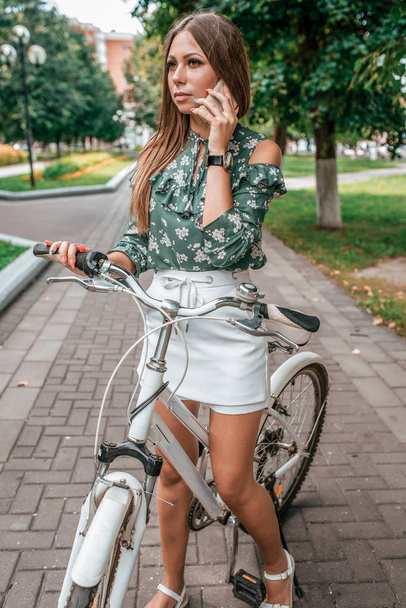 Beautiful girl brunette woman standing in park on a bicycle. Calls by phone in the summer in the city on the background of the road from tiles and green trees. - Fotografie, Obrázek