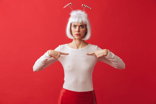 Image of a displeased woman wearing christmas carnival costume over red wall pointing to herself. - Foto, afbeelding
