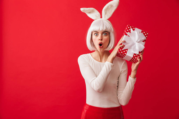 Cheerful young girl wearing Christmas bunny ears standing isolated over red background, holding gift box - Photo, image
