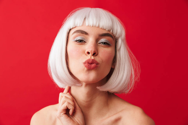 Изображение молодой счастливой женщины, одетой в карнавальный парик, позирующей изолированно на фоне красной стены, дующей поцелуи
. - Фото, изображение