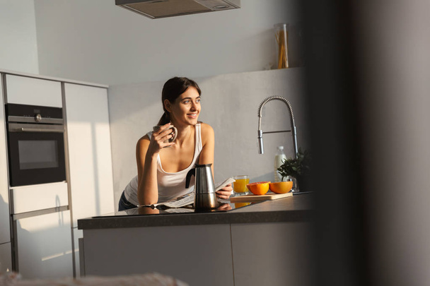 Happy young girl standing at the kitchen in the morning, having cup of coffee - 写真・画像