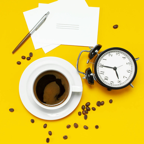 flache Tasse schwarzer Kaffee, Kaffeebohnen, schwarzer Wecker, Stifte, weiße Karten auf gelbem Hintergrund Kopierraum von oben. kreatives Konzept Zeit zum Arbeiten, weiblicher Desktop, Kaffeehintergrund. - Foto, Bild