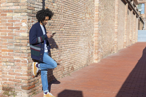 Portrait Young handsome African man using his smartphone with smile while leaning on a bricked wall outdoors in sunny day - Photo, Image