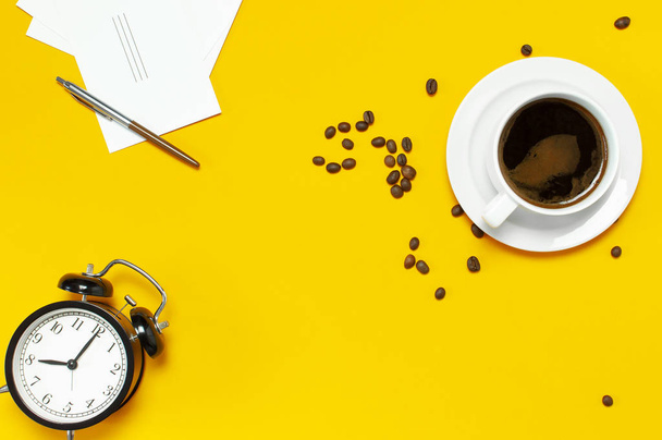 Flat lay cup black coffee, coffee beans, black alarm clock, pens, white cards on yellow background top view copy space. Creative Concept time to work, female desktop, coffee background. - Photo, Image