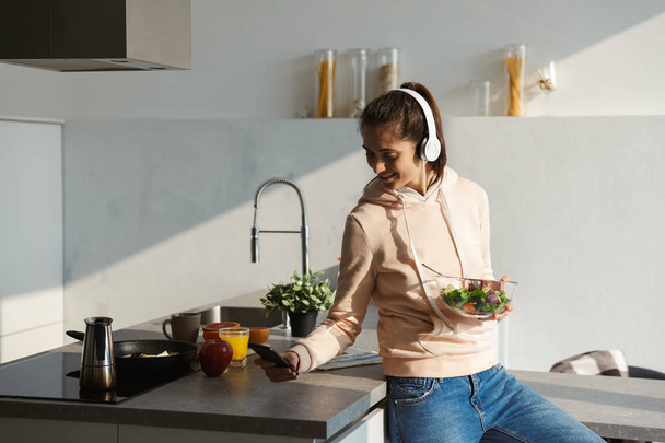Cheerful young girl listening to music with headphones at the kitchen at home, eating salad from a bowl - Photo, image