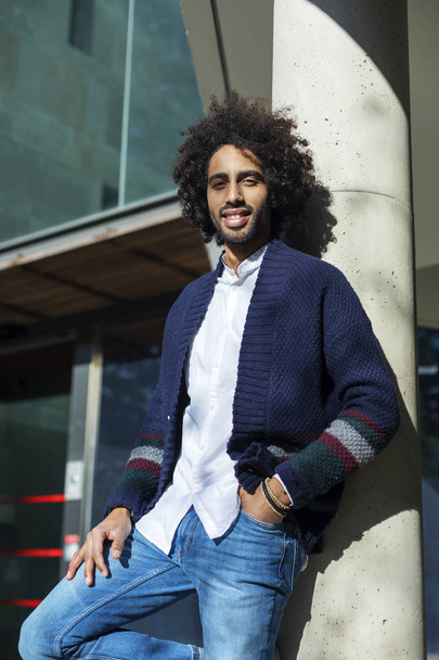 Portrait of handsome Afro American man in casual clothes, looking camera and smiling while leaning on a column, standing outdoors - Foto, Imagen