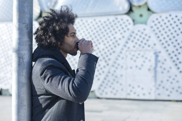 Side view of a Hipster afro man standing and drinking a takeaway coffee, smiling pleasantly, walking on th city street. Happy carefree handsome guy in eye-wear. Millennial generation - Foto, immagini