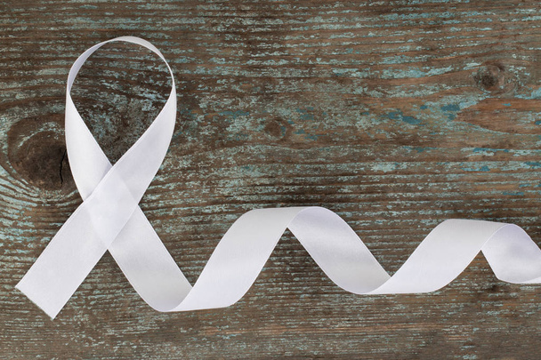 white ribbon on the wooden background with copy space. White or light pearl color ribbon for raising awareness on Lung cancer, Bone cancer, Multiple Sclerosis, Severe Combined Immune Deficiency Disease (SCID) and Newborn Screening  symbol. - Photo, Image