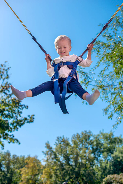 Emotionele portret van het jongetje met veiligheidsgordels op een attractie "Trampoline". Hij met vreugde zweefvliegen in lucht tegen de achtergrond van de blauwe hemel  - Foto, afbeelding