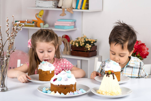 Studio shot van kleine kinderen, meisje en jongen, zitten aan een tafel en eten Pasen taarten.   - Foto, afbeelding