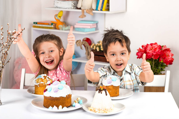 Studio shot of small children, girl and boy, sitting at a table with Easter cakes.  They have a festive mood - Photo, Image