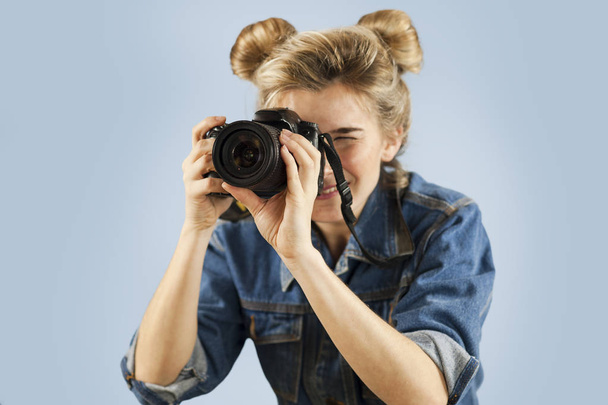 Attractive young woman with a photo camera in her hand in the studio on an isolated blue background. The concept of taking pictures and video - Foto, afbeelding
