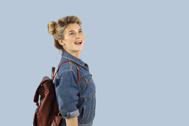 Щаслива красива студентка в джинсовій куртці з рюкзаком на плечах в студії на синьому фоні. Концепція освіти та навчання
. - Фото, зображення