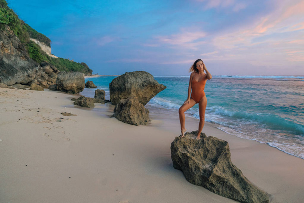 Chica delgada en traje de baño de pie en piedra grande en el fondo de la playa salvaje de hermosa puesta de sol
 - Foto, imagen
