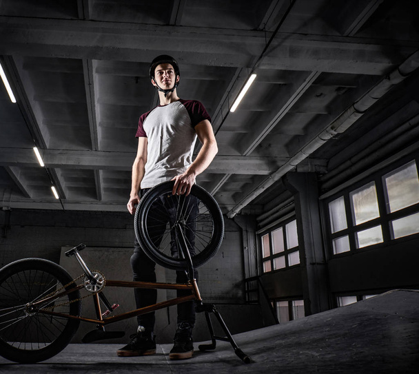 Ganzkörperporträt eines BMX-Profis mit Schutzhelm und seinem Fahrrad, das in einem Skatepark im Innenraum steht - Foto, Bild
