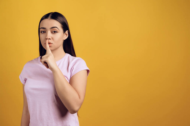 Young girl shows gesture quietly, putting index finger to lips. Isolated on yellow background. Studio portrait. - Фото, зображення