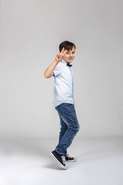 Volledige lengte shot van een lachende jongen draagt een blauw shirt en spijkerbroek wandelen en het tonen van een gebaar van de overwinning op grijze achtergrond in de studio - Foto, afbeelding
