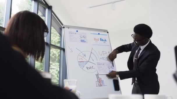 Young businessman conducts presentation using whiteboard on which shows the graphs buying and selling company, afro-american businessman gives presentation in office - Záběry, video