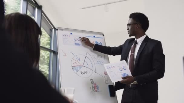 Young businessman conducts presentation using whiteboard on which shows the graphs buying and selling company, afro-american businessman gives presentation in office - Video