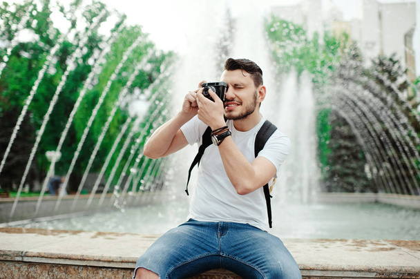 Handsome young man taking photo in park. Stylish boy in a white t-shirt, with bagpack taking photos. - Zdjęcie, obraz
