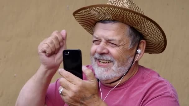 Pozytywne Brodaty starszy człowiek w słomkowy kapelusz, słuchać muzyki z telefonu komórkowego podczas siedzenia przed mur glinie - Materiał filmowy, wideo