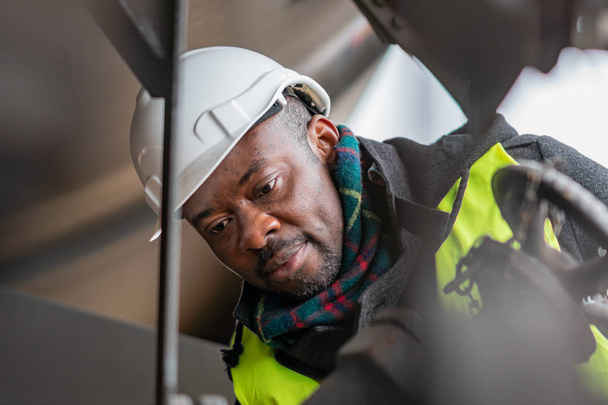 Афроамериканський механік, одягнений в захисне обладнання (шолом і куртка), перевіряє і перевіряє зубчастий поїзд. - Фото, зображення