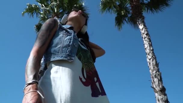 tattooed arrogant brunette taken from downwards against tall swinging in the wind palm trees at the summer sunshine - Materiał filmowy, wideo