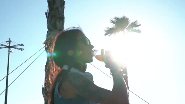 Asian tattooed girl drinking juice in the sunshine and shadow of the palm tree - Materiał filmowy, wideo