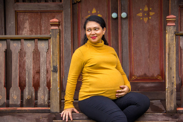 Беременная женщина азиатского происхождения сидит перед старым традиционным деревенским домом. Поза бокового вида
 - Фото, изображение
