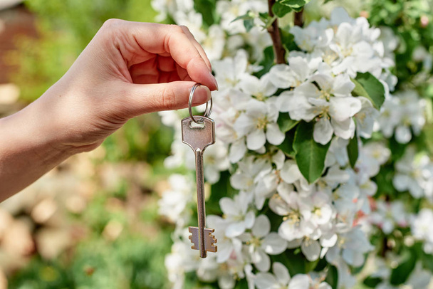 Woman holding in hand house key with blooming apple tree on background, copy space. Spring, real estate agent, new home, buying house property, environment and ecology concept - Photo, Image