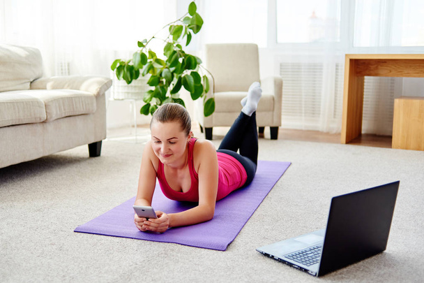 Happy smiling young woman in sportwear relaxing on mat on floor and using mobile cellphone at home, copy space. Full length body. Sport, healthy lifestyle concept  - Foto, Imagen