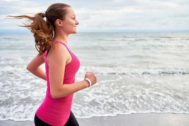Happy fitness woman runner running at seaside, copy space. Girl working out on beach at summer morning, side view. Healthy lifestyle concept - Photo, Image