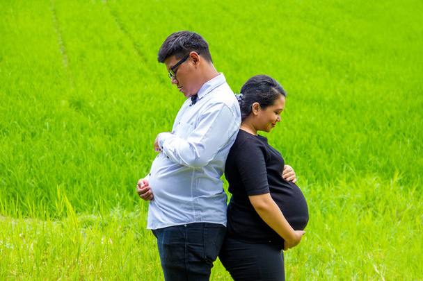 Ázsiai férj és terhes feleség tartja a tummies, természetes legelők háttér áll vissza, vissza, - Fotó, kép