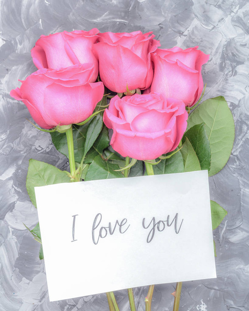 Ystävänpäivän konsepti. Punaisia ruusuja harmaalla betonitaustalla ja postikortti, johon on kaiverrettu "rakastan sinua".
. - Valokuva, kuva