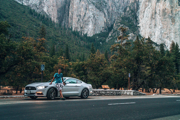 Yosemite National Park, USA. September 10, 2018. Young man standing by the beautiful white Ford Mustang GT parked in the heart of the Yosemite National park with amazing half dome cliffs around it. - Fotografie, Obrázek