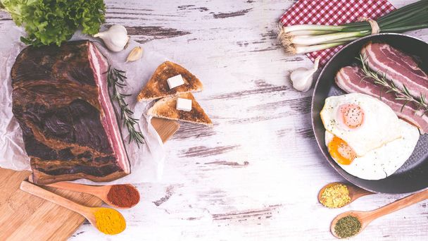 Αυγά τηγανιτά σε τηγάνι, μπέικον, τοστ, σαλάτα και μπαχαρικά σε ξύλινα ρουστίκ φόντο. Πολύ νόστιμο φαγητό. Το top view. Αντιγραφή και επικόλληση - Φωτογραφία, εικόνα