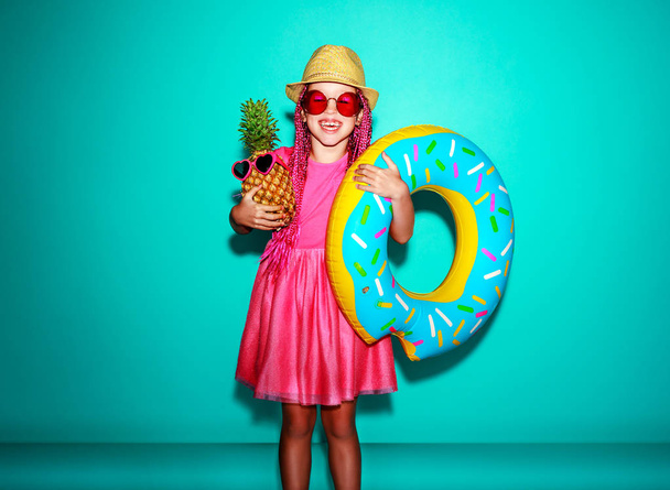 Grappige gelukkig kind meisje in roze zomerjurk met ananas en zwemmen cirkel lachen op blauwe pagina - Foto, afbeelding