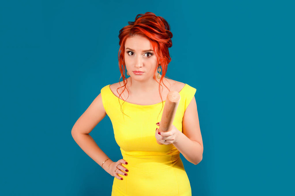 Angry upset frustrated woman holding roll dough stick threatening someone. Caucasian person in yellow dress and retro retro vintage redhead hairstyle on blue studio background. Parenting and relations - Photo, Image